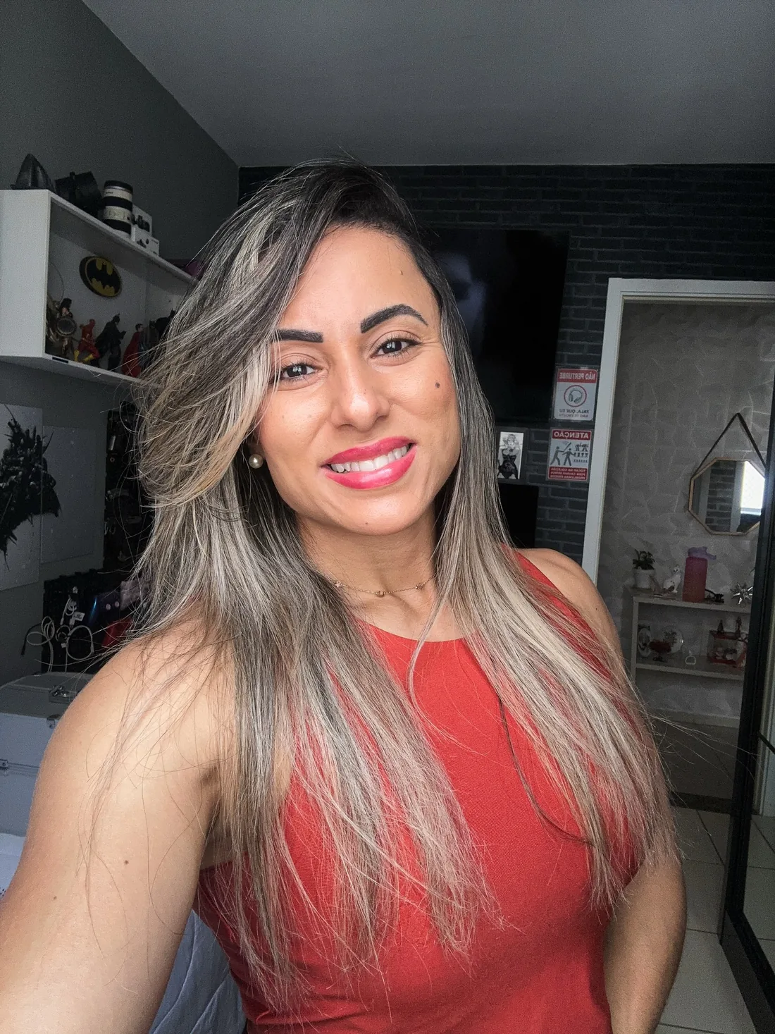 Retrato de Monique Ribeiro sorrindo, cabelo longo com mechas loiras e regata vermelha