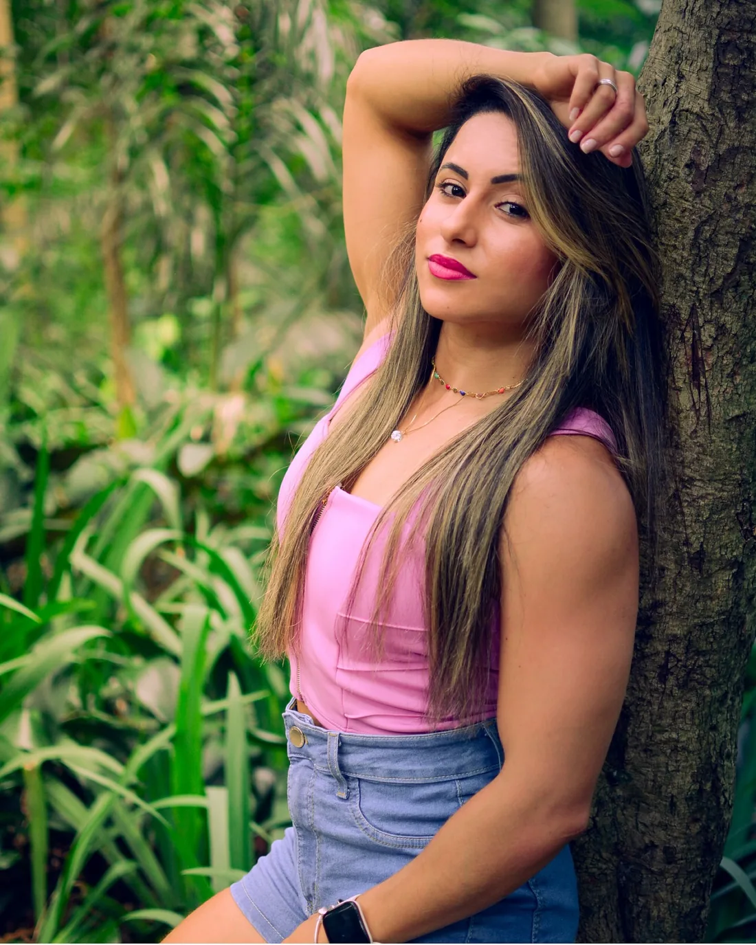Monique Ribeiro de regata rosa com o braço sobre a cabeça, encostada numa árvore em ensaio urbano