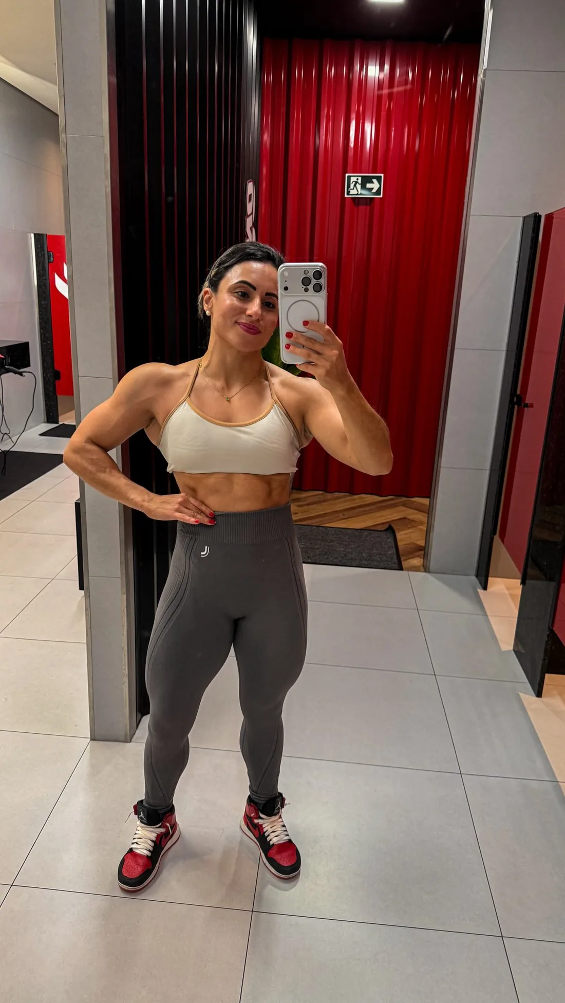 Monique Ribeiro de top branco e legging cinza em selfie no espelho do vestiário